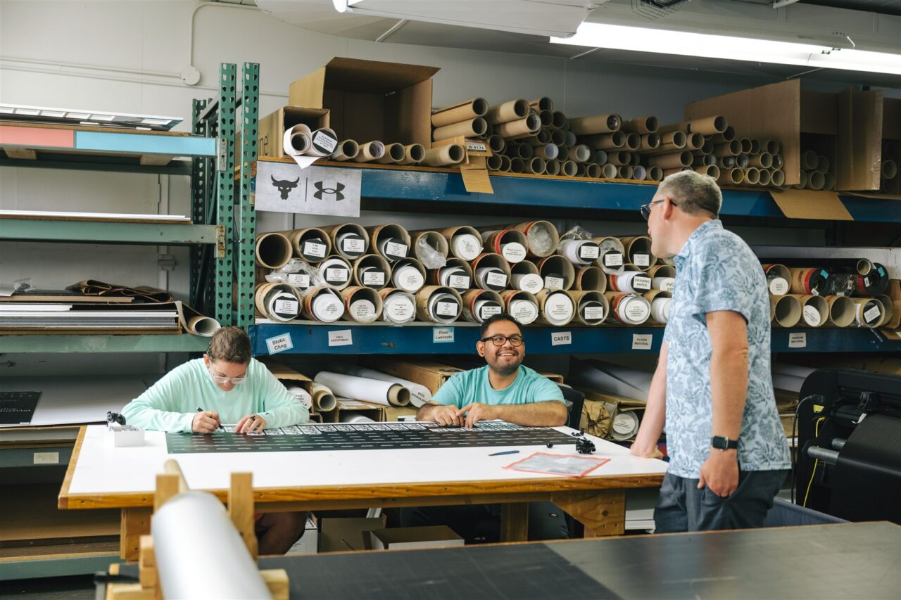 Three Morel Ink team members showcase their wide format print capabilities with a drafting table and shelves full of large banners