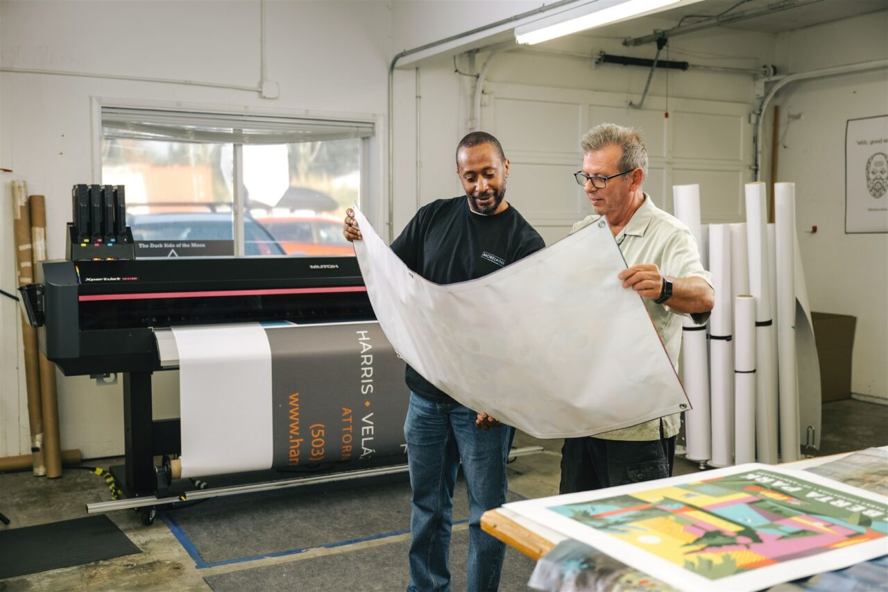 Two Morel Ink team members showcase their wide format printing capabilities, posing with a large banner in front of their equipment